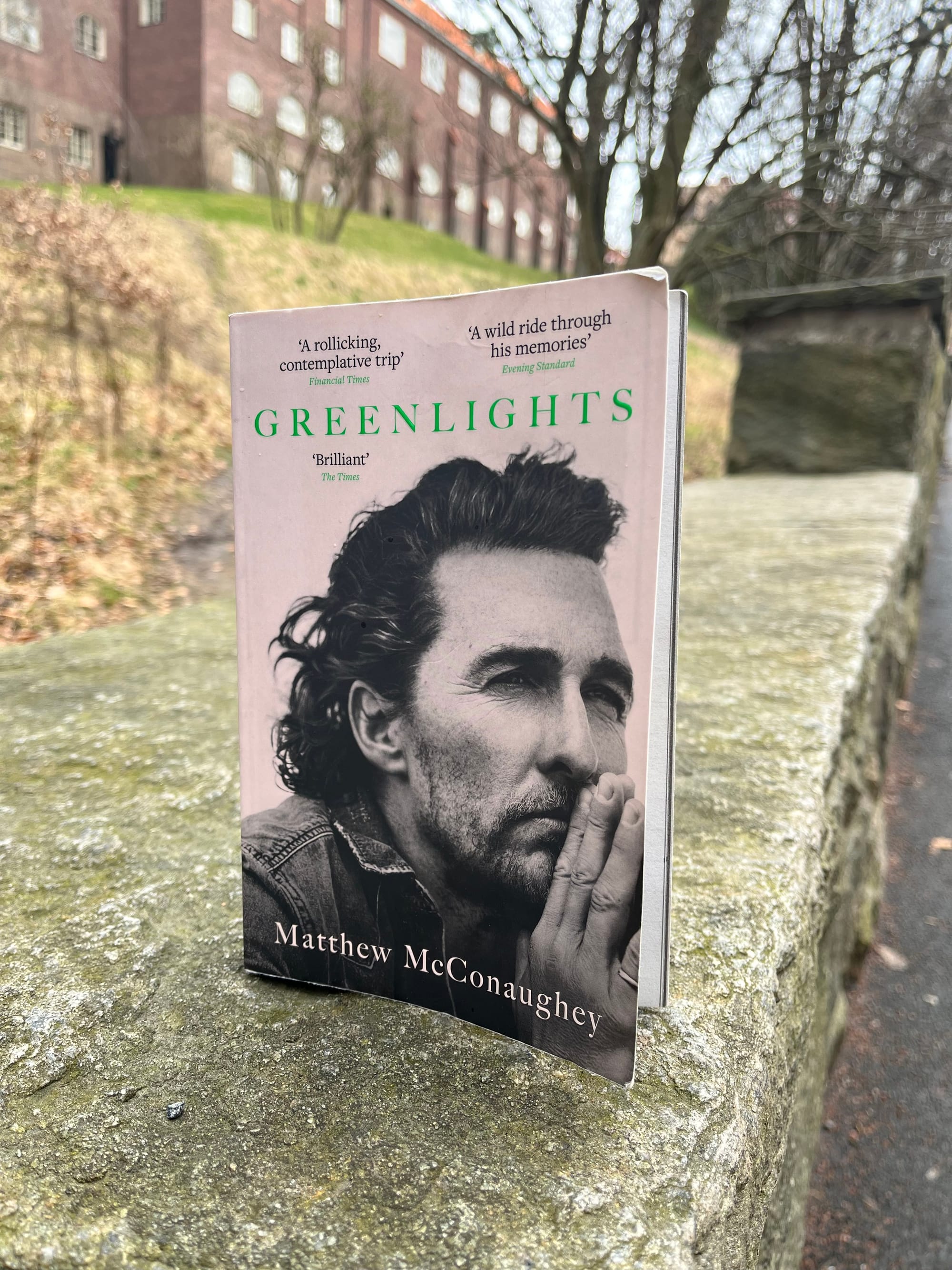The book "Greenlights" on top of a stone wall with a hill and a brick house in the background